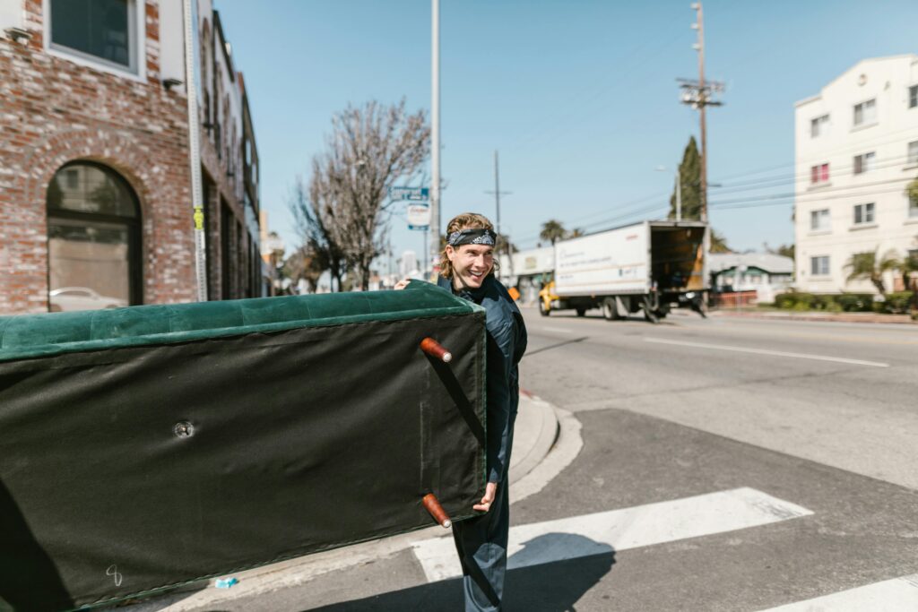 A cheerful man carries a large couch across a sunny urban street during a move.